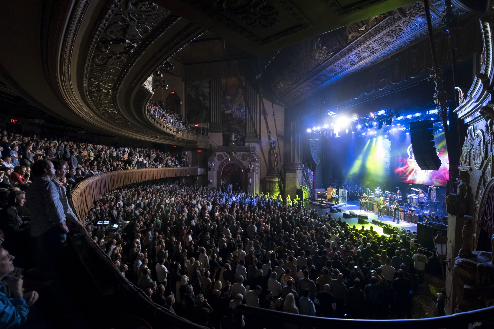 The Beacon Theatre is shown with its beautiful golden statues and art and engraved walls. The seats are red and the curtains of the stage are red as well.