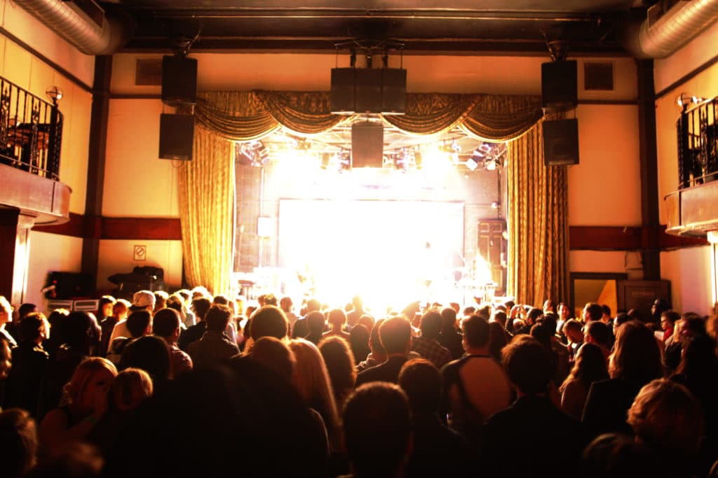 The Bowery Ballroom is shown with curtains around the stage and bright light shining from the stage. The crowd is shown in front of the stage.
