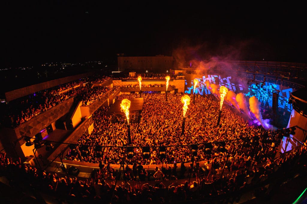 The Brooklyn Mirage and Avant Gardner is shown from a bird's eye view with a crowd amongst flames bursting upward. The stage has blue lighting and billows of smoke shooting upward.