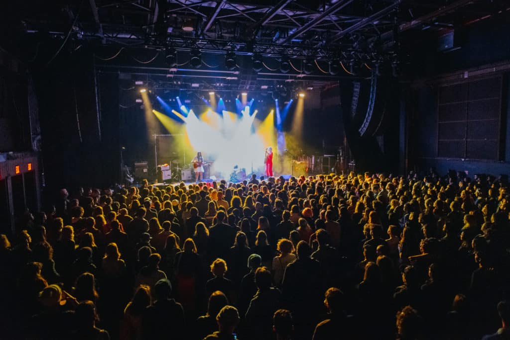 Brooklyn Steel is shown with blue and yellow lighting and performers on stage. The crowd is standing in front in the otherwise dark room.