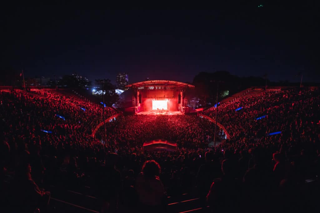 Forest Hills Venue is shown with red light coming from the stage as an elevated view from the top of the amphitheatre shows a full crowd watching a performance against the night sky.
