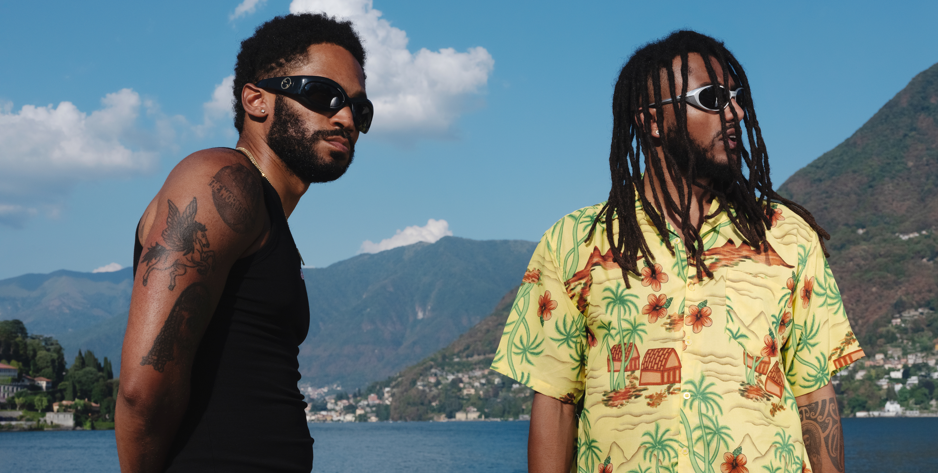 KAYTRANADA and Aminé are shown in front of an ocean and rolling hills with blue skies. KAYTRANADA wears a black tank top and sunglasses while Aminé wears a yellow buttoned shirt with houses and palm trees on it as well as grey sunglasses.