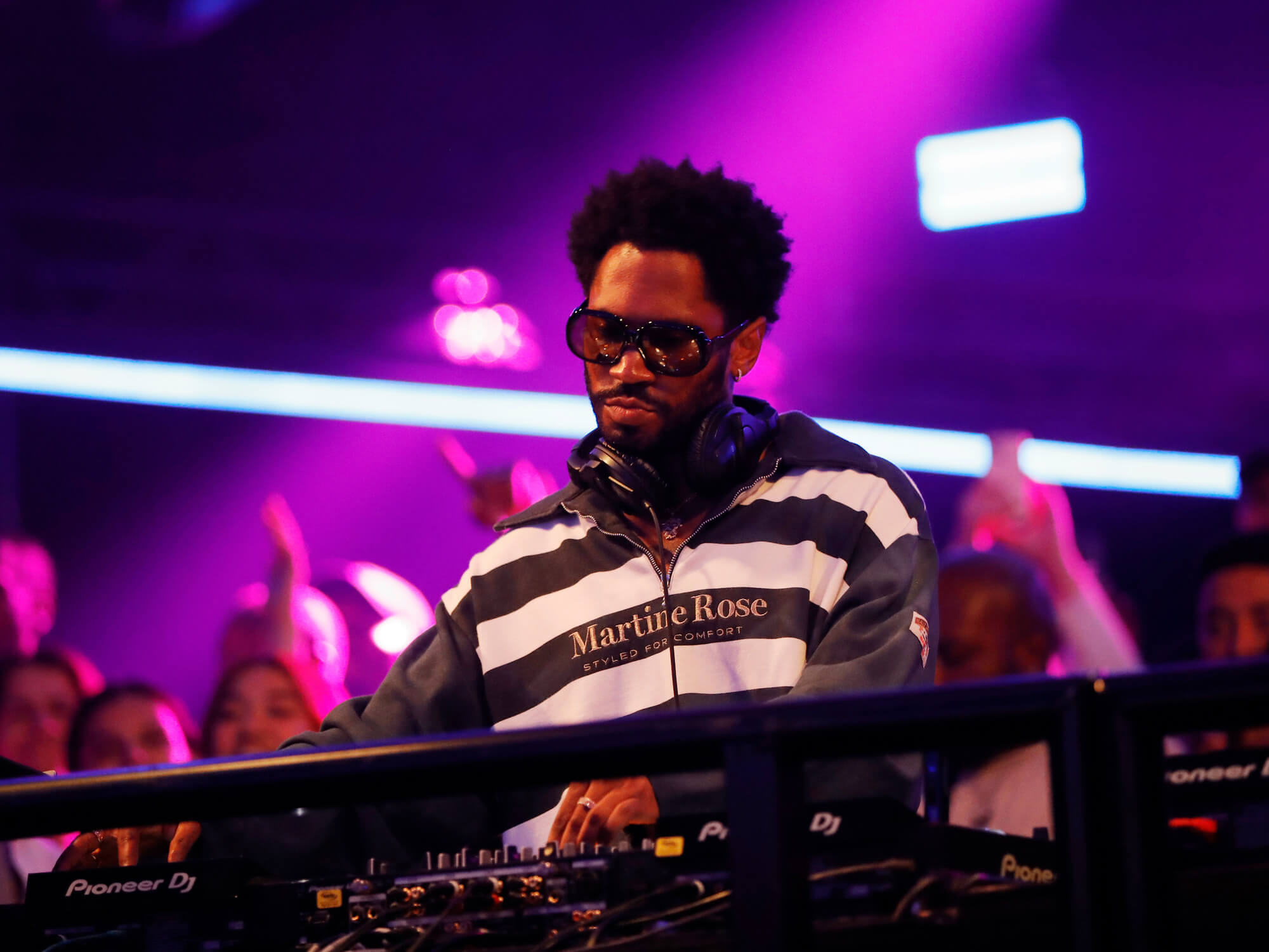 KAYTRANADA is shown performing in a club in front of DJ equipment with a crowd behind him and purple lighting inside.