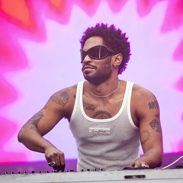 KAYTRANADA is shown performing with DJ equipment wearing a white tank top and black goggle sunglasses on a pink, purple, and orange star-pattern background.