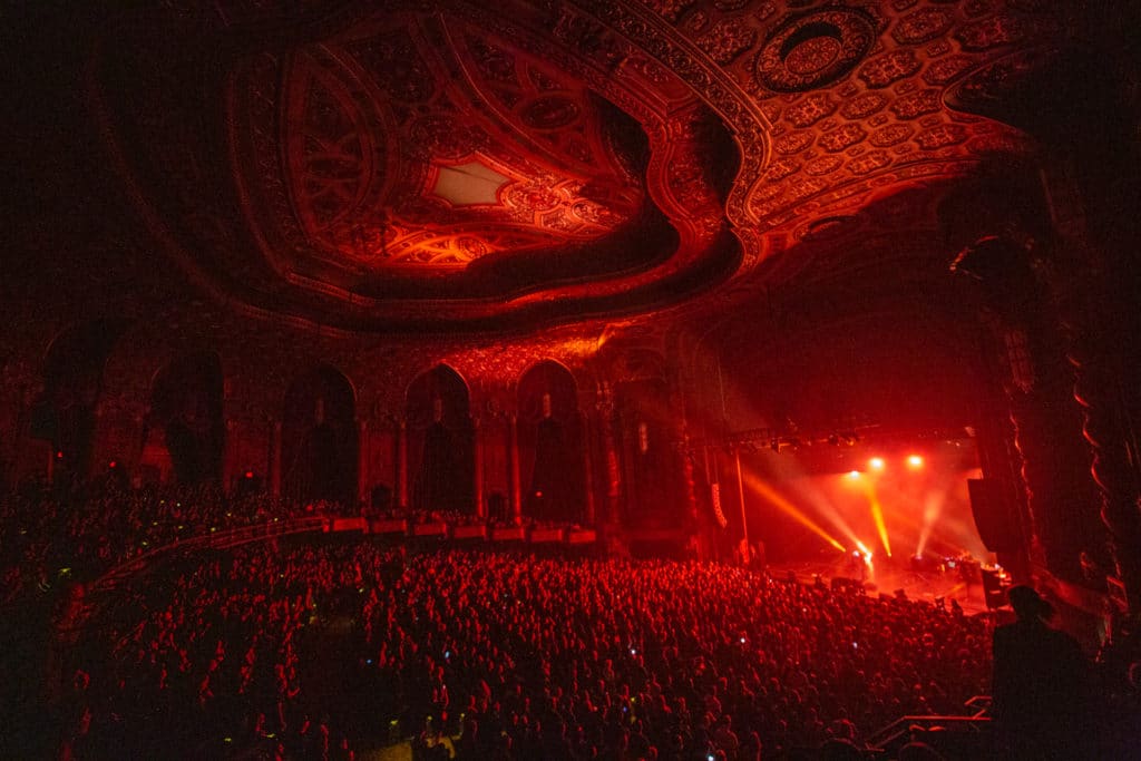 Kings Theatre is shown lit by red lights from an elevated view with many people in the crowd in front of a stage and ornate engravings on the ceilings and walls