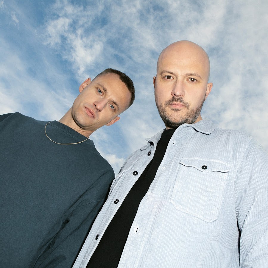 the duo of Overmono stand above the camera facing down with a blue sky and clouds behind them. one is wearing a turquoise shirt with a gold chain and the other is wearing a black shirt and blue corduroy button up outer shirt.