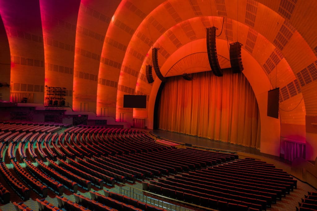 Radio City Music Hall is shown empty with orange lighting cascading over the stage and empty rows seats. There are curtains and speakers on stage, and the ceiling shows ridges as it rounds higher.