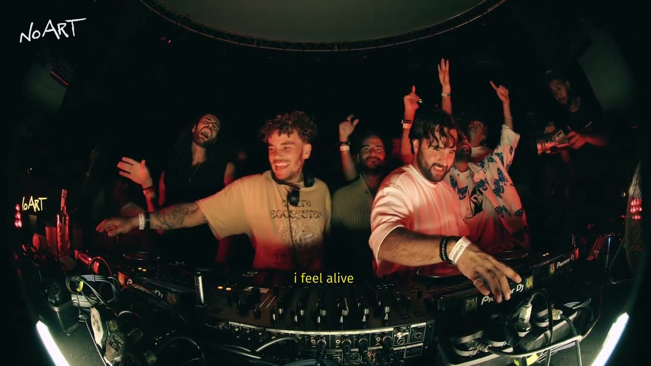 two DJ's are pictured in front of their mixing equipment with 3 people behind them. Red lighting surrounds them and they have smiles on their faces.
